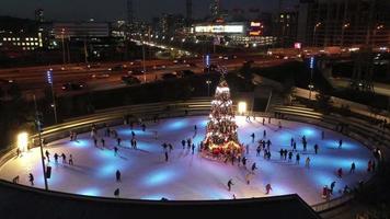 People ice skate around a Christmas tree in a skating rink at night with light show video