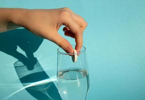 Against a blue background, a hand drops a dissolving fizzy aspirin tablet into a glass of water photo