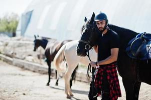 El hombre árabe de barba alta usa casco negro con caballo árabe. foto