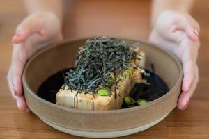 cierre un tofu frío con salsa de soja dashi, cubriendo algas y edamame en un cuenco de barro de estilo japonés con la mano de una mujer sosteniendo un cuenco. foto