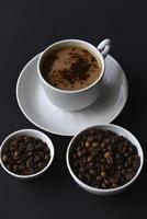 delicioso café en una taza de porcelana con platillo y granos de café. café en una taza y frijoles sobre un fondo negro. foto
