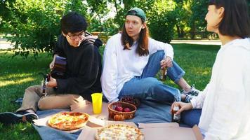 des amis sont assis sur une couverture dans le parc et parlent avec des boissons et de la guitare video