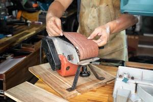 carpintero que usa delantal e instala o quita la correa abrasiva de la lijadora de banda en tabel de madera en el taller, el fabricante de bricolaje y el concepto de carpintería. enfoque selectivo foto