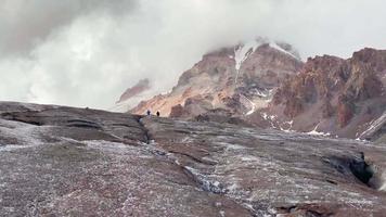 vista posterior vista inspiradora caminata de dos excursionistas masculinos en un terreno glaciar helado con el pico de la montaña kazbek en el parque nacional kazbegi. glaciar gergeti kazbek ruta de senderismo video