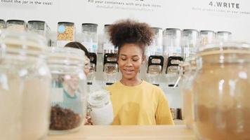 Two young female customers choose and shop for organic products in refill store, zero-waste grocery, plastic-free, and reusable containers, Thai texts on wall mean fill container with any bulk item. video