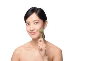 foto de estudio de una atractiva joven asiática usando un rodillo de jade en la cara contra un fondo blanco.