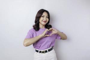 An attractive young Asian woman wearing a lilac purple t-shirt feels happy and a romantic shapes heart gesture expresses tender feelings photo