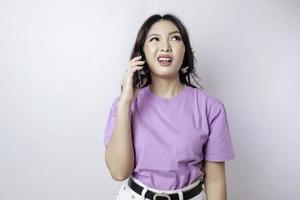 Confused Asian woman wearing a lilac purple t-shirt while picking up a phone call, isolated by a white background photo