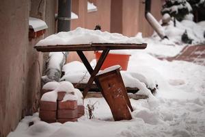 Close up of a small table and bench in a yard with red bricks pile standing near and covered in snow photo