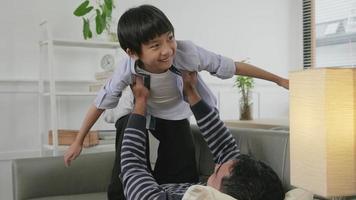 sian tailandese famiglia insieme, padre bugie giù e divertimento giochi con figlio di sollevamento, diffusione braccia piace volante aereo su vivente camera divano, contento tempo libero volte, bello fine settimana, benessere domestico stile di vita. video