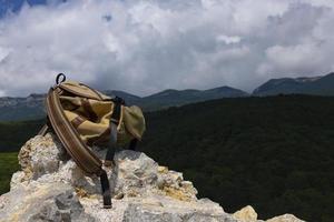 A brown-green backpack lies on a large gray stone on a rock against the backdrop of green mountains. photo