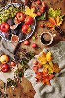 plato con higos, manzanas y uvas y taza de café sobre fondo de madera con un cálido suéter, bufanda, hojas de otoño y manzanas. fondo de otoño, vista superior. foto