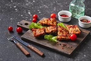 Grilled baked pork ribs with spices and vegetables on wooden cutting board on dark background. American food concept. photo
