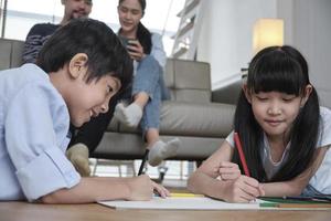 Asian Thai siblings are lying on living room floor, drawing homework with colored pencils together, parents leisurely relax on a sofa, lovely happy weekend activity, and domestic wellbeing lifestyle. photo