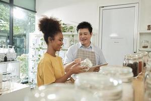 Asian male shopkeeper describes natural organic products to woman customer in refill store, zero-waste and plastic-free grocery, eco environment-friendly, sustainable lifestyles with a reusable shop. photo