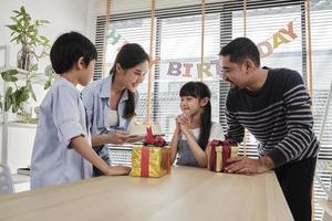 Happy Asian Thai family, young daughter is surprised with birthday cake and gift, blows candle, pray and cheerful celebrates party with parents together at dining table, domestic home event lifestyle. photo