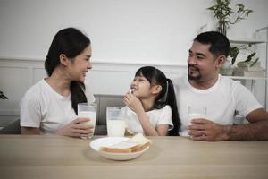 una familia tailandesa asiática saludable, una hija pequeña y padres jóvenes beben leche blanca fresca en un vaso y pan de alegría juntos en una mesa de comedor en la mañana, estilo de vida de comida de desayuno en casa de nutrición de bienestar. foto