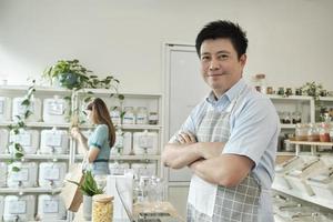 Portrait of Asian male shopkeeper smiles and looks at camera, arms crossed at refill store, natural products, zero-waste grocery, and plastic-free, eco environment-friendly, sustainable lifestyles. photo