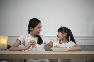 una familia tailandesa asiática saludable, una hija pequeña y una madre joven beben leche blanca fresca en un vaso y pan de alegría juntos en una mesa de comedor por la mañana, bienestar nutrición hogar desayuno comida estilo de vida. foto