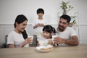 una familia tailandesa asiática saludable, niños pequeños y padres jóvenes beben leche blanca fresca en un vaso y pan de alegría juntos en una mesa de comedor en la mañana, bienestar nutrición hogar desayuno comida estilo de vida. foto