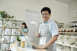 Portrait of happy Asian male shopkeeper smiles looks at camera, and thumbs up at refill store, natural products, zero-waste grocery, and plastic-free, eco environment-friendly, sustainable lifestyles. photo