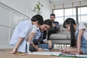 Asian Thai siblings and mum are sitting on living room floor, drawing with colored pencils together, dad leisurely relaxing on a sofa, lovely happy weekend activity, and domestic wellbeing lifestyle. photo