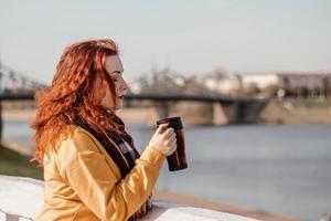 Young red-haired woman in yellow sweater holds thermo mug with coffee in her hands. Enjoy the river view on sunny autumn day. photo
