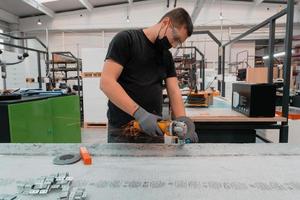 interior de la fábrica de ingeniería de la industria pesada con un trabajador industrial que usa una amoladora angular y corta un tubo de metal. Lleva una máscara en la cara debido a la pandemia del coronavirus foto