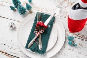 Christmas table setting with white dishware, silverware and red and green decorations on white wooden background. High angle view.
