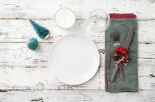 Christmas table setting with white dishware, silverware and red and green decorations on white wooden background. Top view. photo