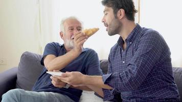 glückliche familie mit senior vater und sohn essen brot mit leckeren zusammen mit fröhlichen auf dem sofa, junge mann gespräch mit älteren menschen auf der couch im wohnzimmer, lebensstil und beziehungskonzept. video