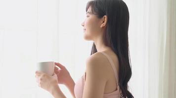 bela jovem asiática feliz e confiante bebendo café com janelas frescas e olhando em casa, estilo de vida garota asiática relaxar no quarto em casa para o conceito de bem-estar e saúde. video