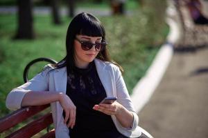 Cute Woman with Sunglasses and Long Hair is Using Mobile Device Enjoying Sunbeams and Warm Day Outdoors. Portrait of Smiling Brunette with Smartphone in the Park.