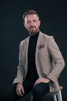 Portrait of adult businessman wearing trendy suit and sitting in modern studio on stylish chair against the black background photo