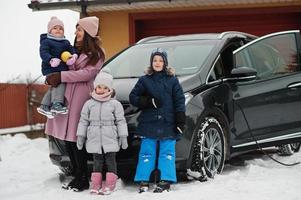 joven madre con hijos cargando un coche eléctrico en el patio de su casa en invierno. foto