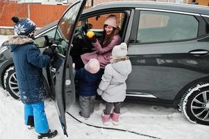 Young mother with kids charging electric car in the yard of her house at winter. photo