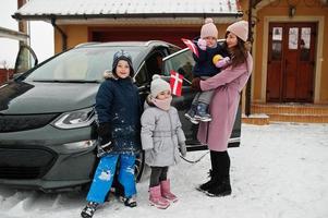 la joven madre danesa con hijos sostiene banderas de dinamarca y carga un coche eléctrico en el patio de su casa en invierno. foto