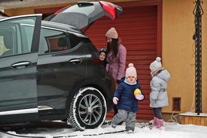 Young woman with children hold eco bags and charging electric car with open trunk in the yard of her house . photo