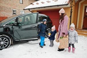 Young woman with kids hold eco bags and charging electric car in the yard of her house . photo