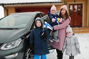 Young finnish mother with kids hold Finland flags and charging electric car in the yard of her house at winter. photo