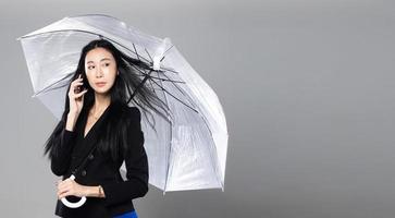 Asian Transgender Woman with long black straight hair, wind blow throw in the Air. Female hold phone and umbrella against wind storm, feeling fashion sensual sexy, gray background isolated copy space photo