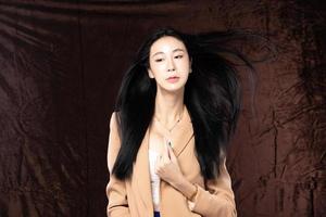 Haft body portrait of beautiful transgender 20s asian woman express feeling smiling confidently on brown background. Business Female smart look blazer has wind blow throw her hair in air, copy space photo