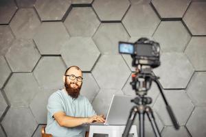 creación de contenidos para redes sociales. hombre barbudo filmando un video de sí mismo usando una cámara en un trípode. tecnología moderna y concepto de trabajo independiente de blogs. foto