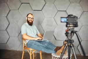 creación de contenidos para redes sociales. hombre barbudo filmando un video de sí mismo usando una cámara en un trípode. tecnología moderna y concepto de trabajo independiente de blogs. foto