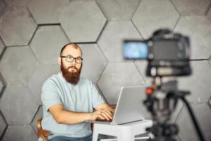 creación de contenidos para redes sociales. hombre barbudo filmando un video de sí mismo usando una cámara en un trípode. tecnología moderna y concepto de trabajo independiente de blogs. foto