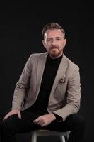 Portrait of adult businessman wearing trendy suit and sitting in modern studio on stylish chair against the black background photo