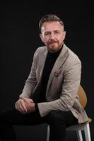 Portrait of adult businessman wearing trendy suit and sitting in modern studio on stylish chair against the black background photo