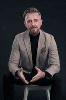 Portrait of adult businessman wearing trendy suit and sitting in modern studio on stylish chair against the black background photo