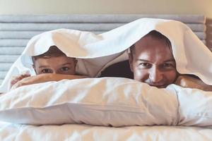 Happy father and son covered with sheets lying down on bed. photo