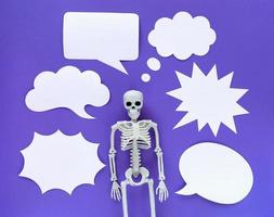 Skeleton on violet background with lots of white blank paper speech bubbles. Anatomical plastic model human skeleton with variety of emotions. Empty dialog clouds, place for text. Purple Halloween.